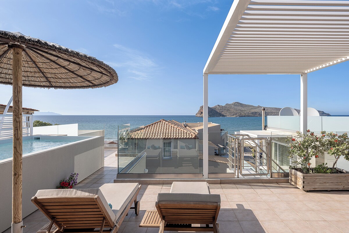 A balcony with a view of the sea, wooden lounge chairs, a glass balustrade, and a pergola.