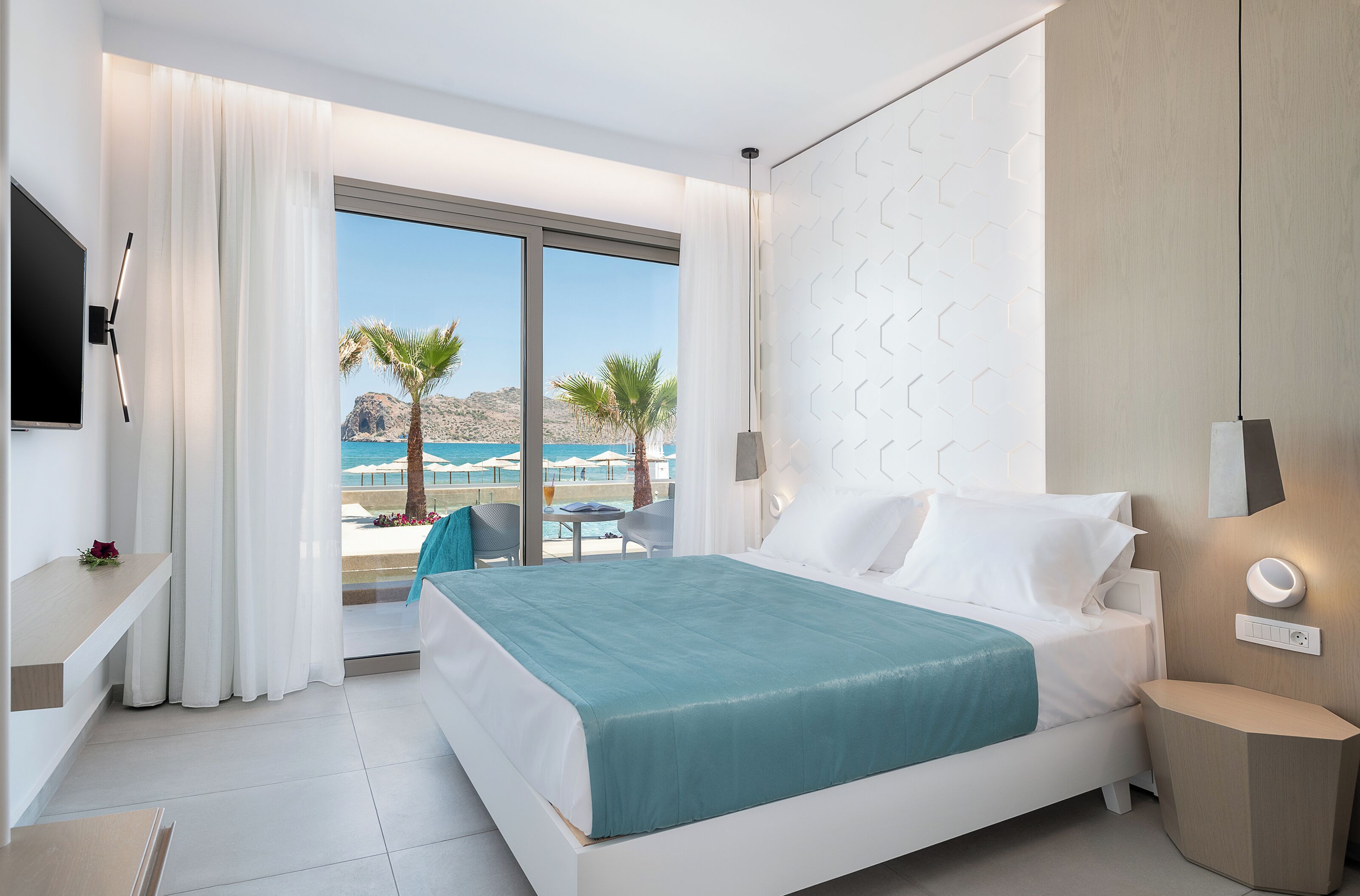 A modern hotel room with a large bed, a TV, a bedside table, and a view of the sea.