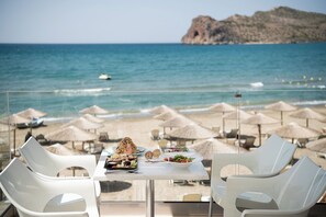 A table with food and two white chairs, overlooking a beach with umbrellas and a rocky island in the distance. - Vergina Beach Resort (Chania)