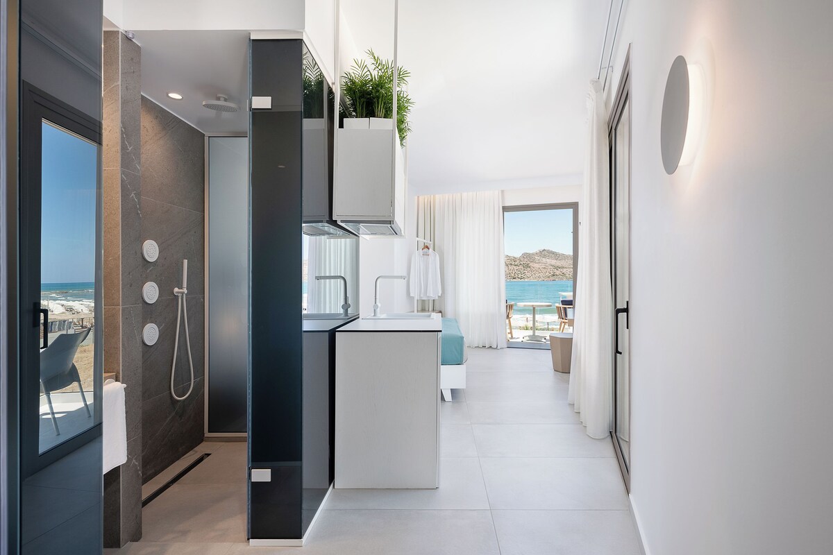 A modern bathroom with a shower, a sink, and a toilet. There is a view of the beach through the window.
