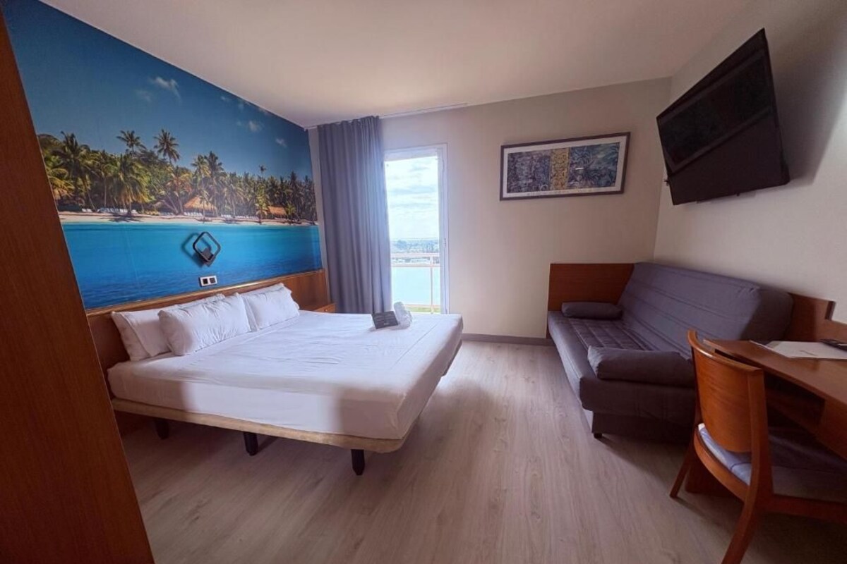 A hotel room with a bed, a sofa, a desk, a TV, and a painting of a beach scene.