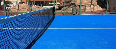 A tennis court with a net, surrounded by a green fence and palm trees.