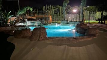 Una piscina nocturna con formaciones rocosas y palmeras.