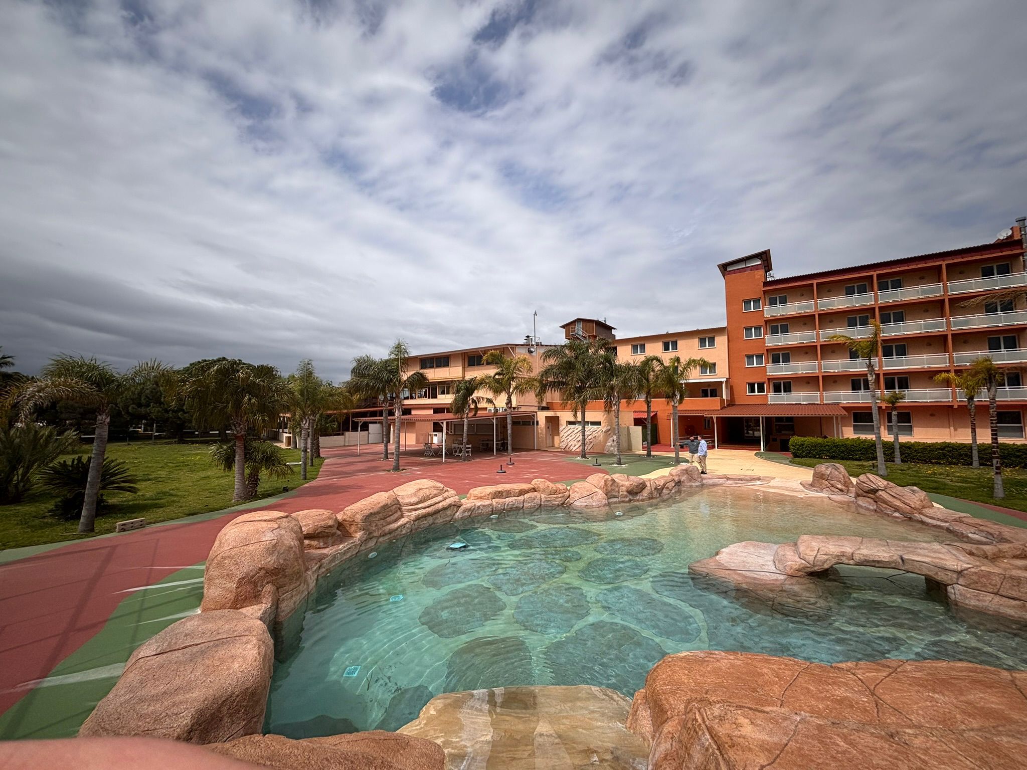 Un hotel con piscina rodeada de rocas, una pista de atletismo roja y palmeras.