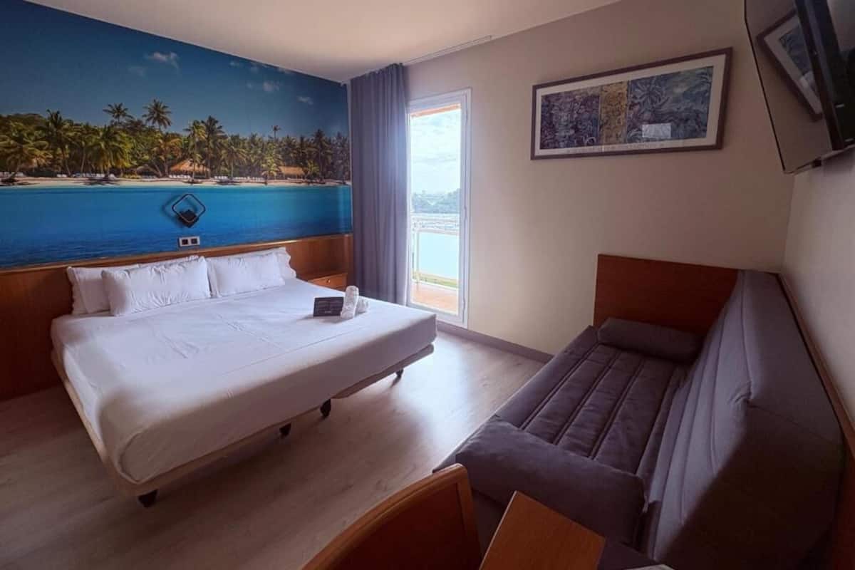 A hotel room with a large bed, a sofa, a painting, and a view of the sea.
