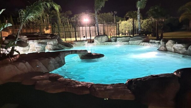 Une piscine éclairée la nuit, avec des rochers sur le bord et des palmiers en arrière-plan.