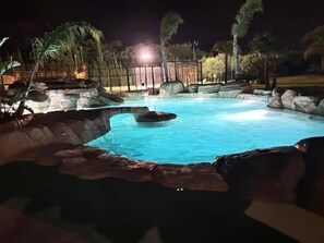 Una piscina nocturna con borde rocoso y palmeras al fondo.