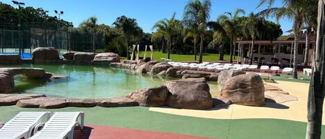 Un área de piscina con rocas grandes, sillas de descanso y palmeras.
