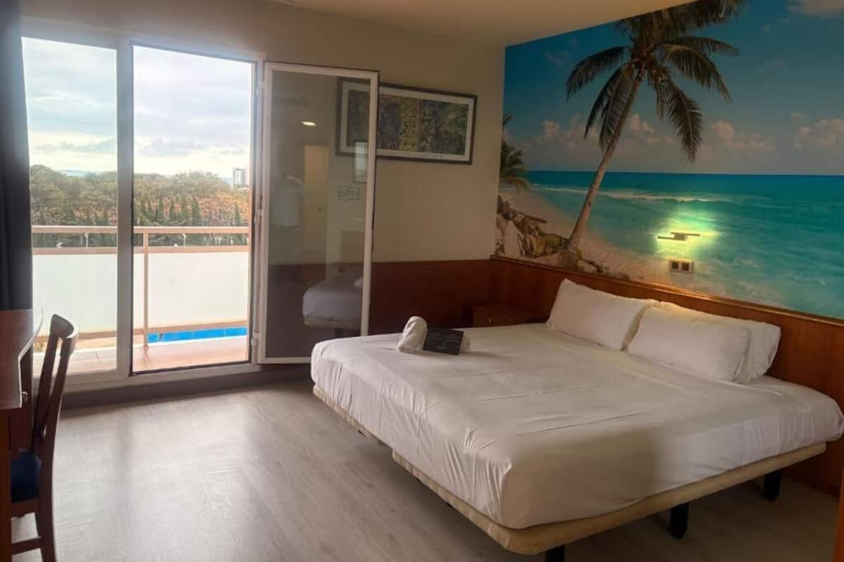 A bedroom with a large bed, a view of the ocean, a balcony, and a painting of a beach scene.
