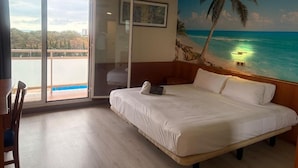Une chambre avec un grand lit, une vue sur l’océan, un balcon et un tableau représentant une scène de plage.