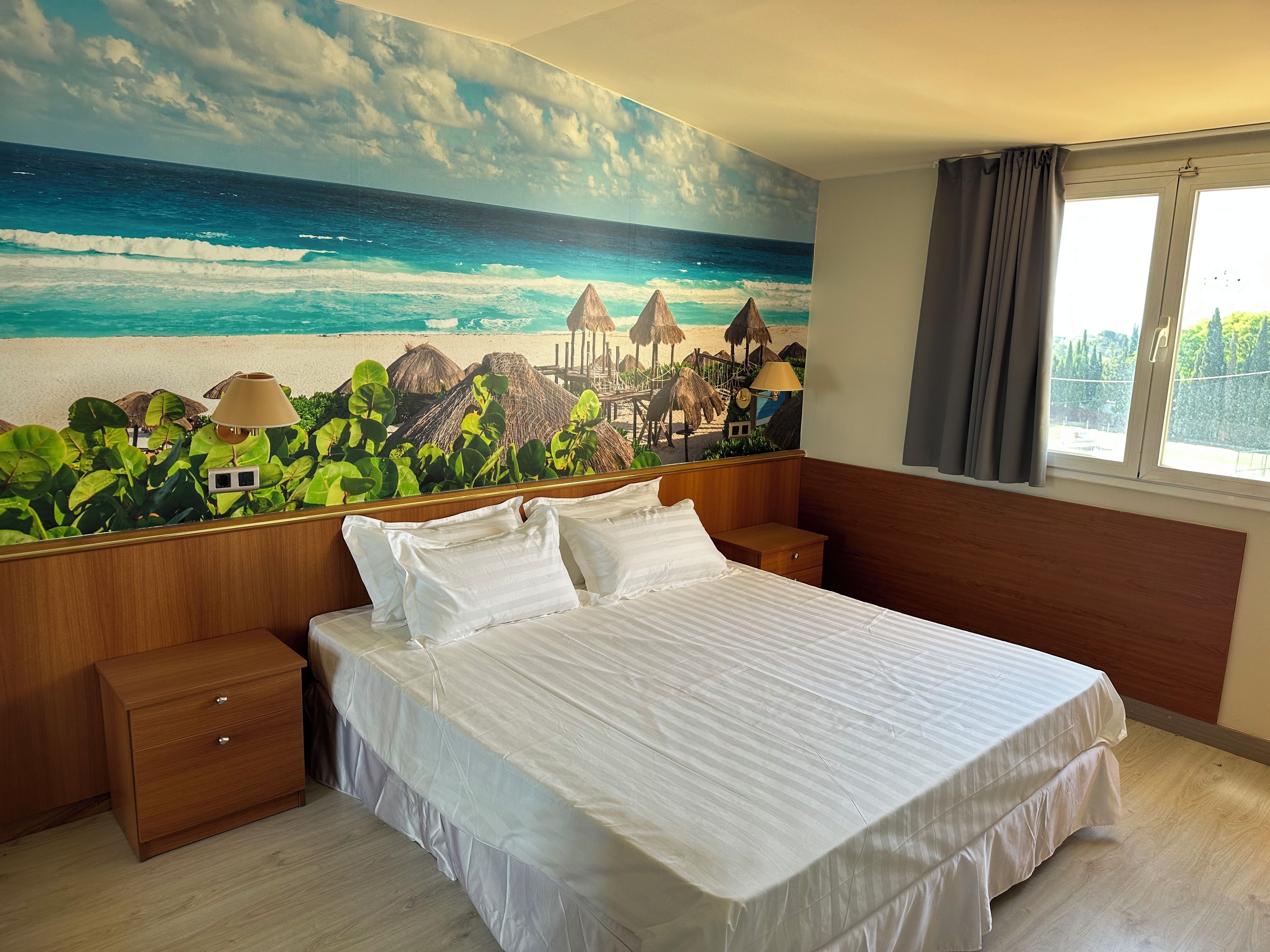 A bedroom with a bed, nightstands, a wooden dresser, and a beach mural on the wall.