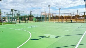 Een buitenterrein voor tennis, met een groene ondergrond, omheind door een hek en voorzien van verlichting.