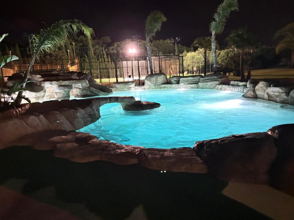 Una piscina nocturna con borde rocoso y palmeras al fondo.
