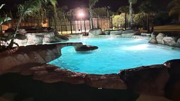 Una piscina nocturna con borde rocoso y palmeras al fondo.