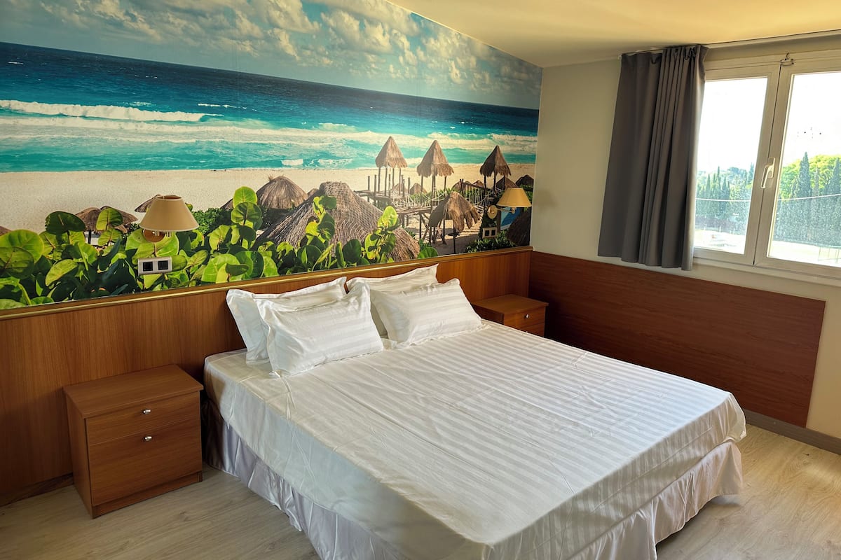 A bedroom with a bed, nightstands, a wooden dresser, and a beach mural on the wall.