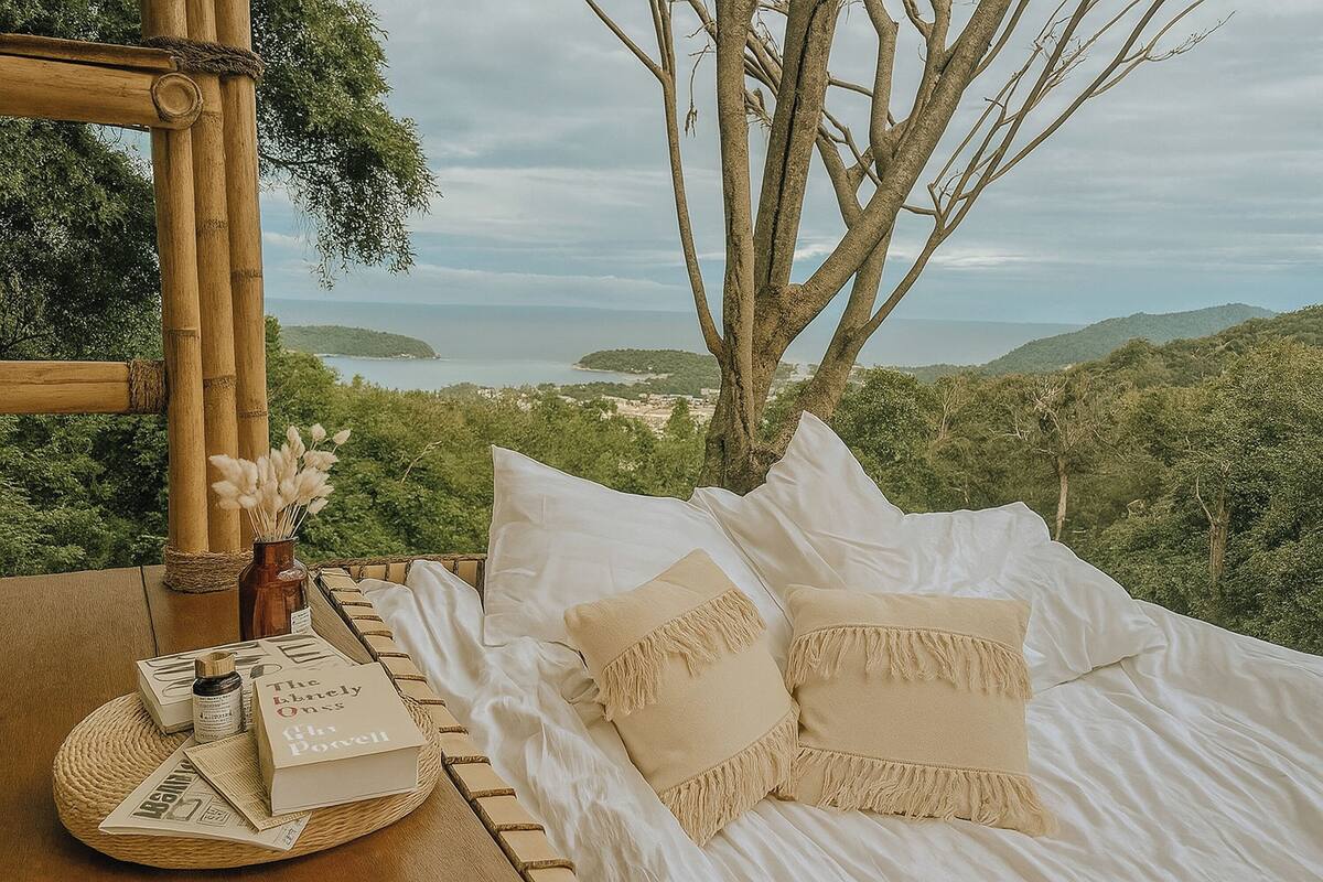 A bed with white linens and beige pillows, a wicker tray with books and a bottle, and a view of the sea and hills.