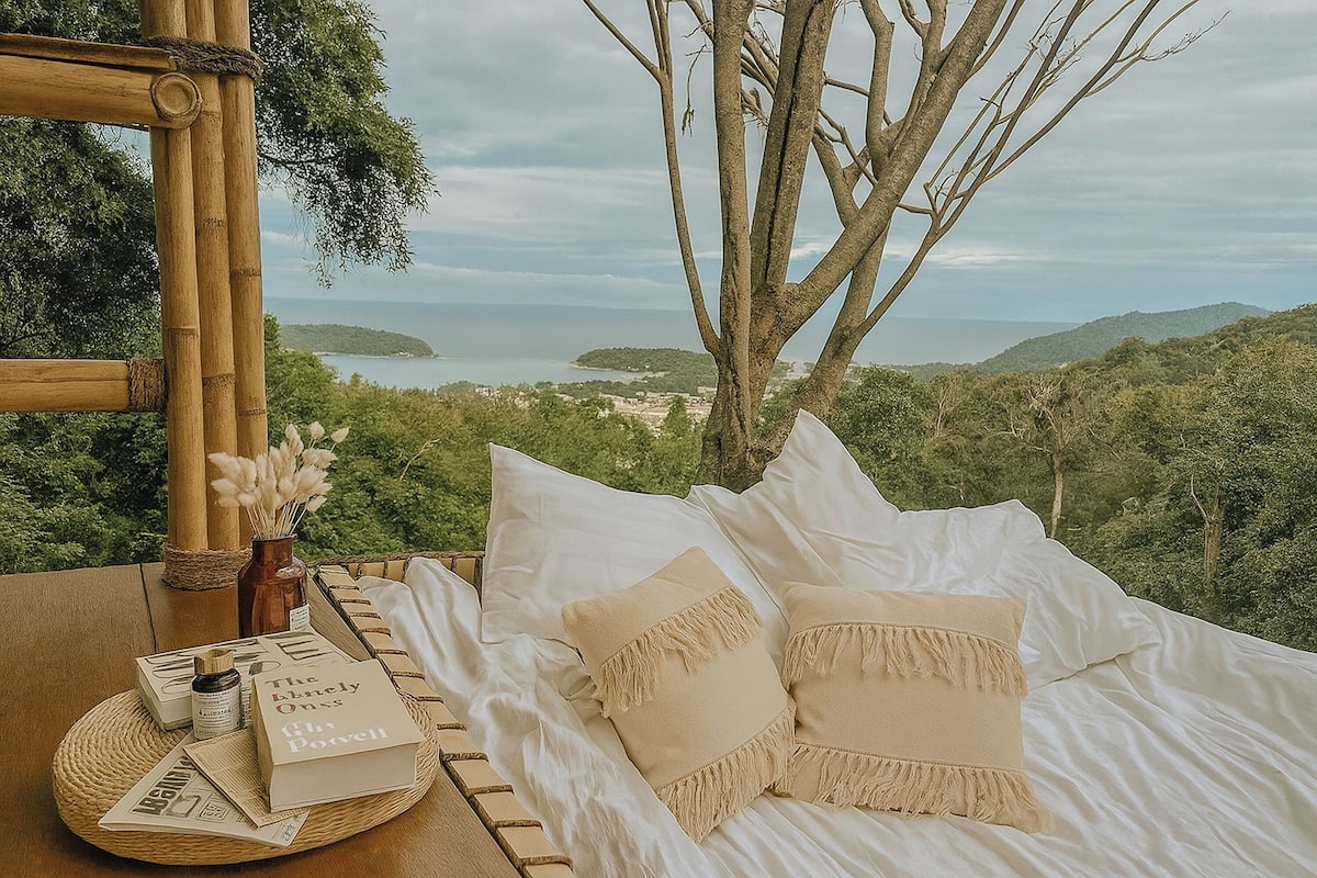 A bed with white linens and beige pillows, a wicker tray with books and a bottle, and a view of the sea and hills.