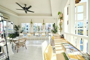 A bright, open-plan living area with a ceiling fan, large windows, and a variety of plants. - Sleepy Nomad Hostel (Malacca City)