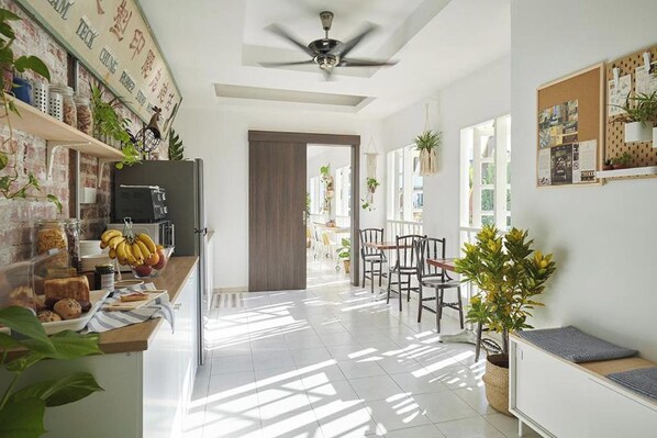 A bright kitchen with a counter displaying food items, a ceiling fan, and a view into the dining area. - Sleepy Nomad Hostel (Malacca City)