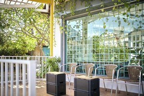 An outdoor seating area with wicker chairs and black stools, a glass window with a grid pattern, and greenery outside. - Sleepy Nomad Hostel (Malacca City)