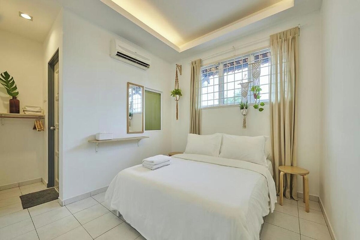 Une chambre d’hôtel avec un lit, un petit tabouret, une fenêtre avec des rideaux, une plante et une étagère avec une serviette.