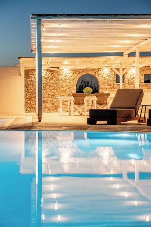 A poolside area with a stone wall, a covered seating area with chairs, and a lounge chair. - Philosophy Of Mykonos (Mykonos)
