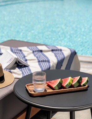 A poolside setting with a table holding a glass of water and slices of watermelon, a striped towel, and a straw hat. - Philosophy Of Mykonos (Mykonos)