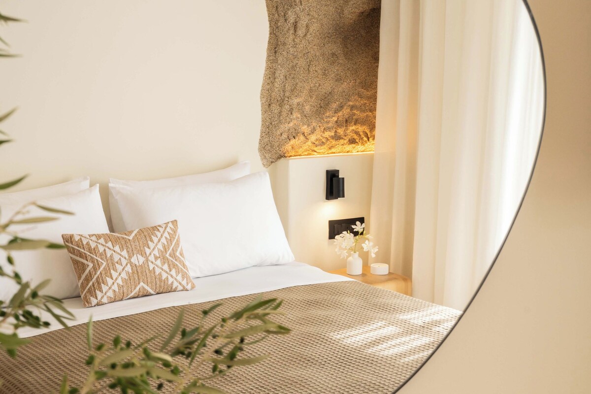 Quarto com cama, almofadas, mesa de cabeceira com um livro e espelho refletindo o aposento.