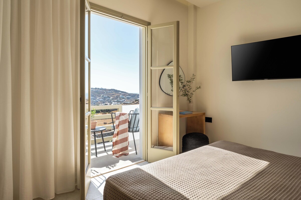 Quarto de hotel com cama, televisão fixada na parede, escrivaninha com cadeira e vista da cidade através de uma grande janela.