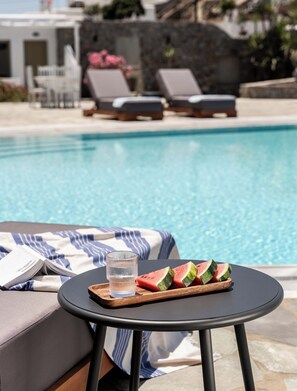 A poolside setting with a table holding a glass of water and watermelon slices, a striped towel, and a book. - Philosophy Of Mykonos (Mykonos)