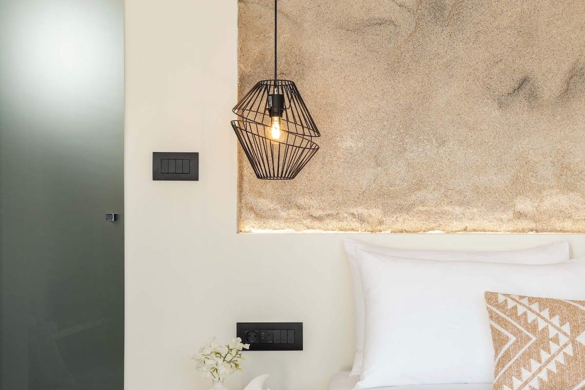 A bedroom with a bed, a wooden nightstand, a wall-mounted light fixture, and a textured wall art piece.