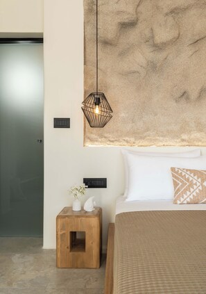 A bedroom with a bed, a wooden nightstand, a wall-mounted light fixture, and a textured wall art piece. - Philosophy Of Mykonos (Mykonos)