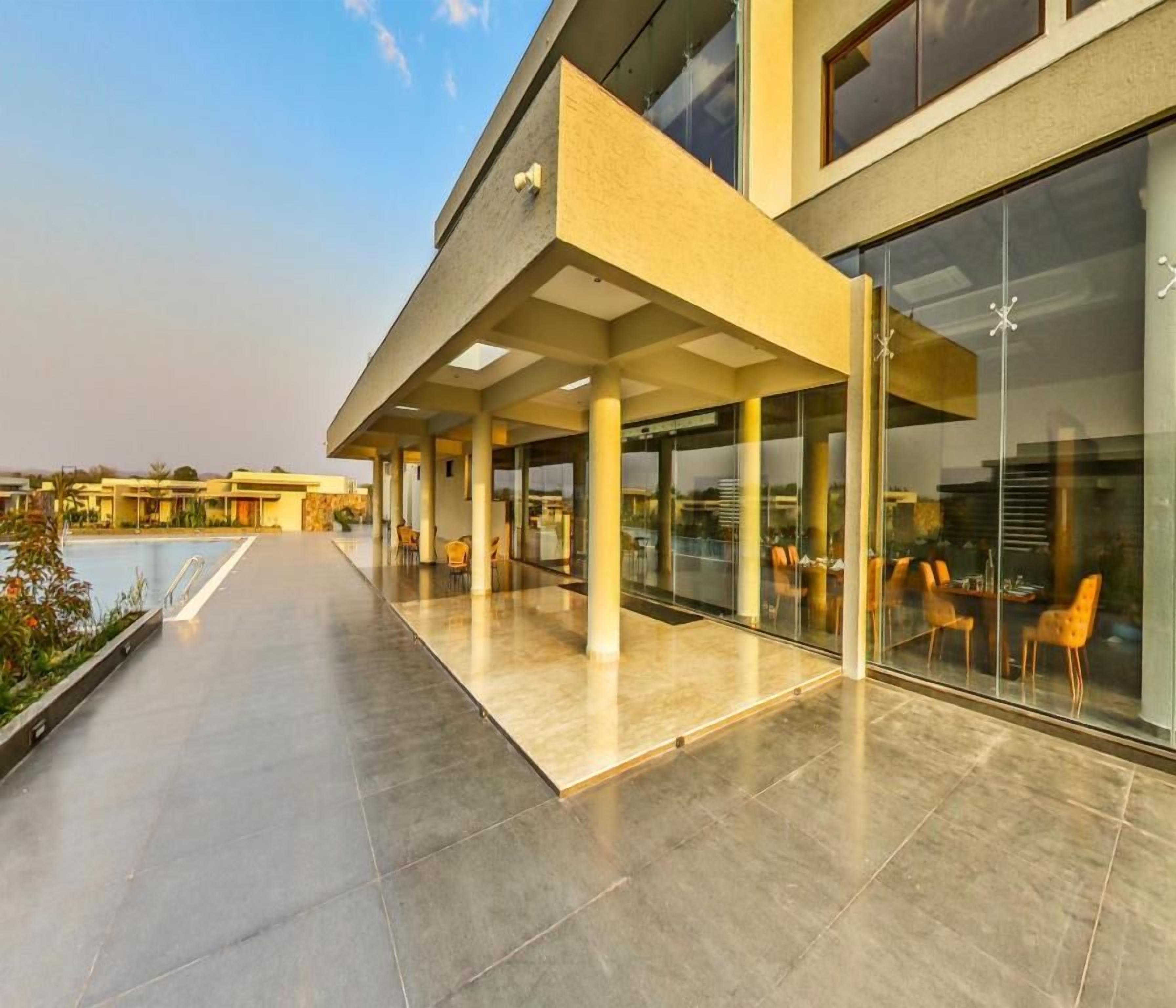 A modern hotel exterior with a covered outdoor seating area, a pool, and large windows.