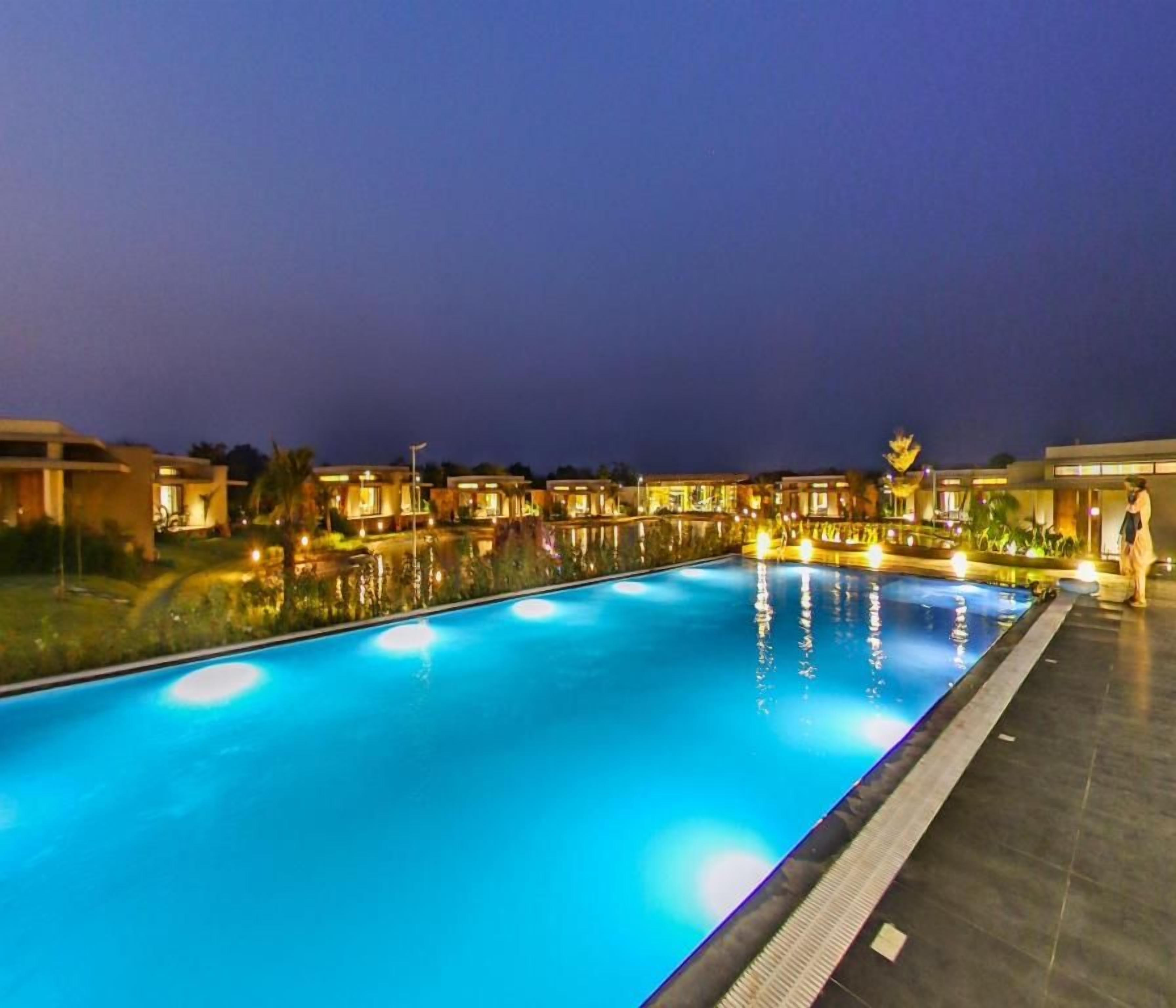 A swimming pool with a tiled deck, surrounded by buildings with warm lighting.