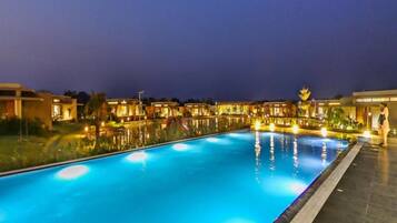 A swimming pool with a tiled deck, surrounded by buildings with warm lighting.