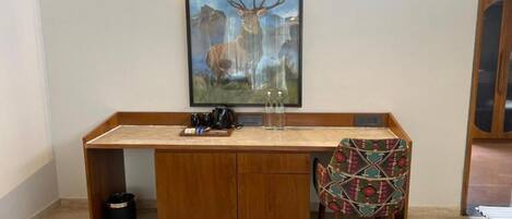 A wooden desk with a framed wildlife painting, a chair with a patterned cushion, and a trash bin.