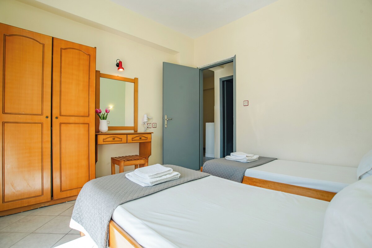 A hotel room with two beds, a wooden wardrobe, a small desk with a mirror, and a door leading to another room.