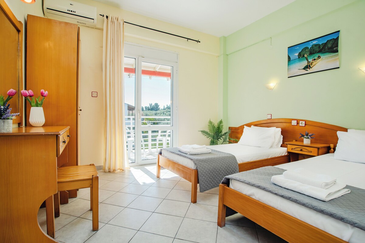 A hotel room with two beds, a desk, a chair, a window with curtains, a potted plant, and a painting on the wall.