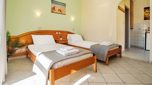 A hotel room with two single beds, a wooden headboard, a nightstand, a wall-mounted lamp, a painting, and a tiled bathroom visible through an archway. - Solena Studios Kavos (Corfu)
