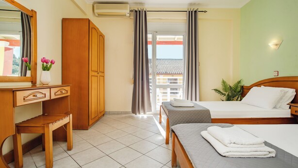A hotel room with a wooden desk, a bed with a grey quilt, a bench, a plant, and a view of a building through a window. - Solena Studios Kavos (Corfu)