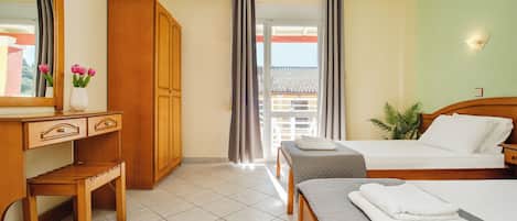 A hotel room with a wooden desk, a bed with a grey quilt, a bench, a plant, and a view of a building through a window.
