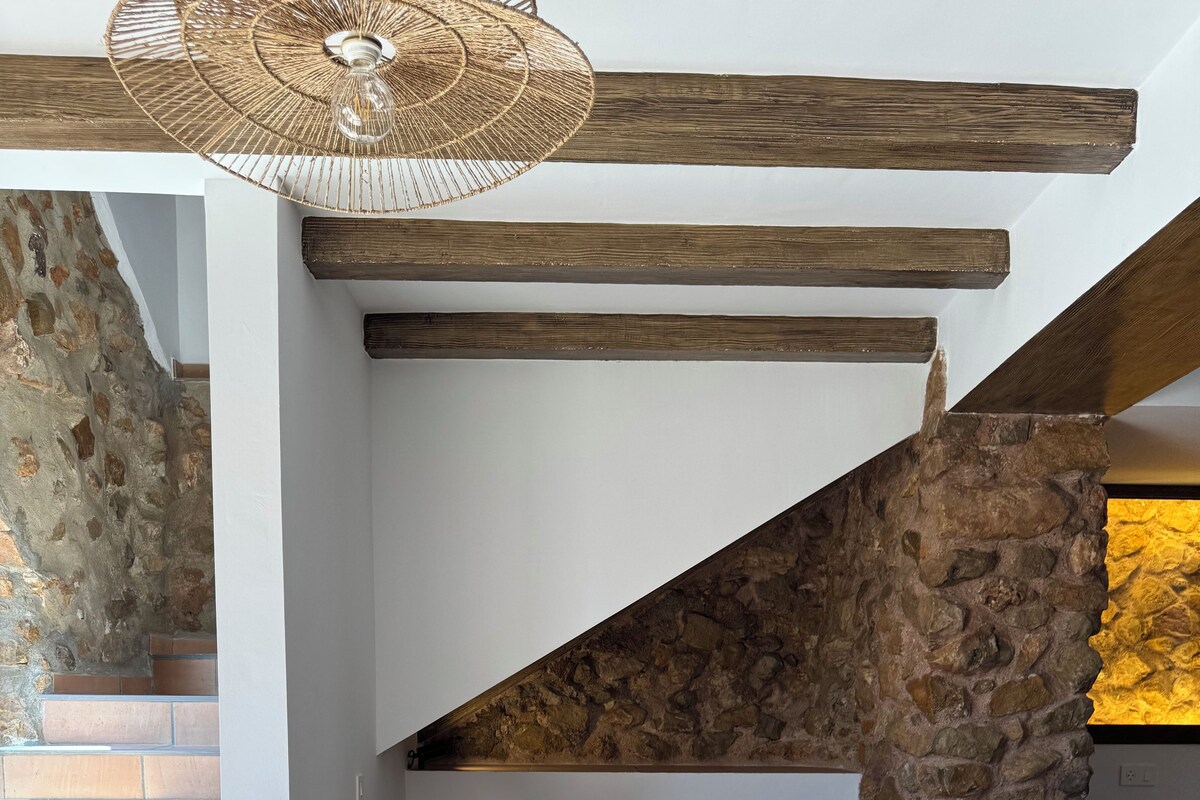 Un comedor con mesa de madera, sillas blancas, una lámpara colgante y una pared de piedra.