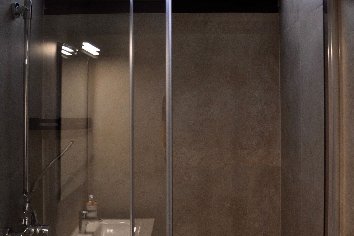 Un baño moderno con ducha envuelta en vidrio, lavamanos a la vista y ventana sobre la ducha.