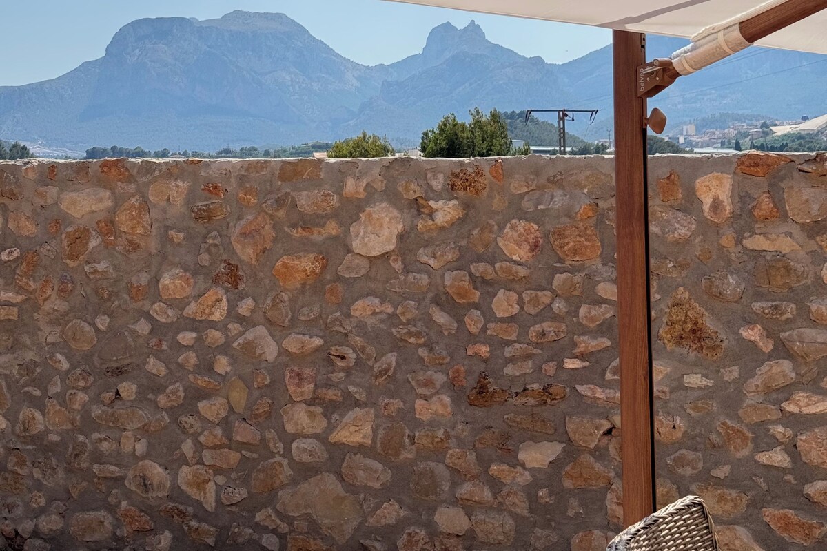 Un muro de piedra, una pérgola de madera y un paisaje montañoso.
