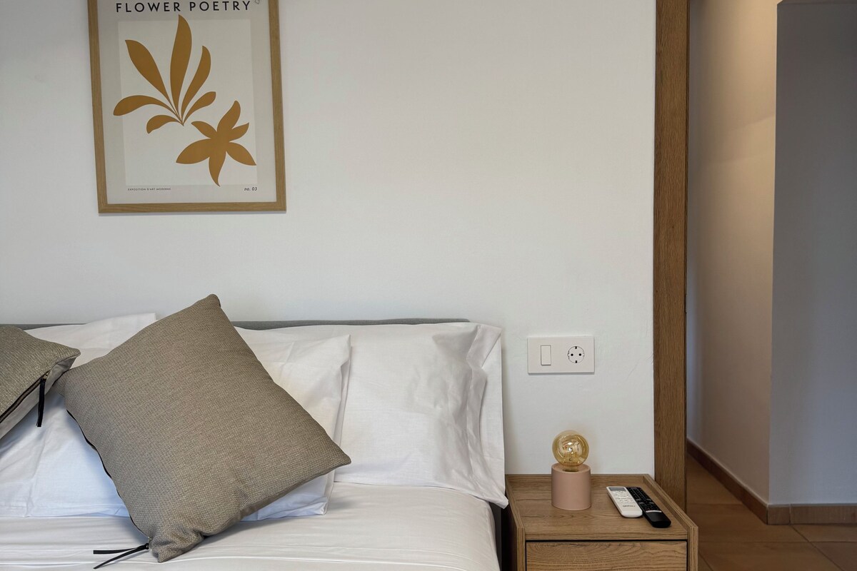 Un dormitorio con cama, almohadas, mesita de noche con lámpara, una obra de arte en la pared y una puerta de madera.