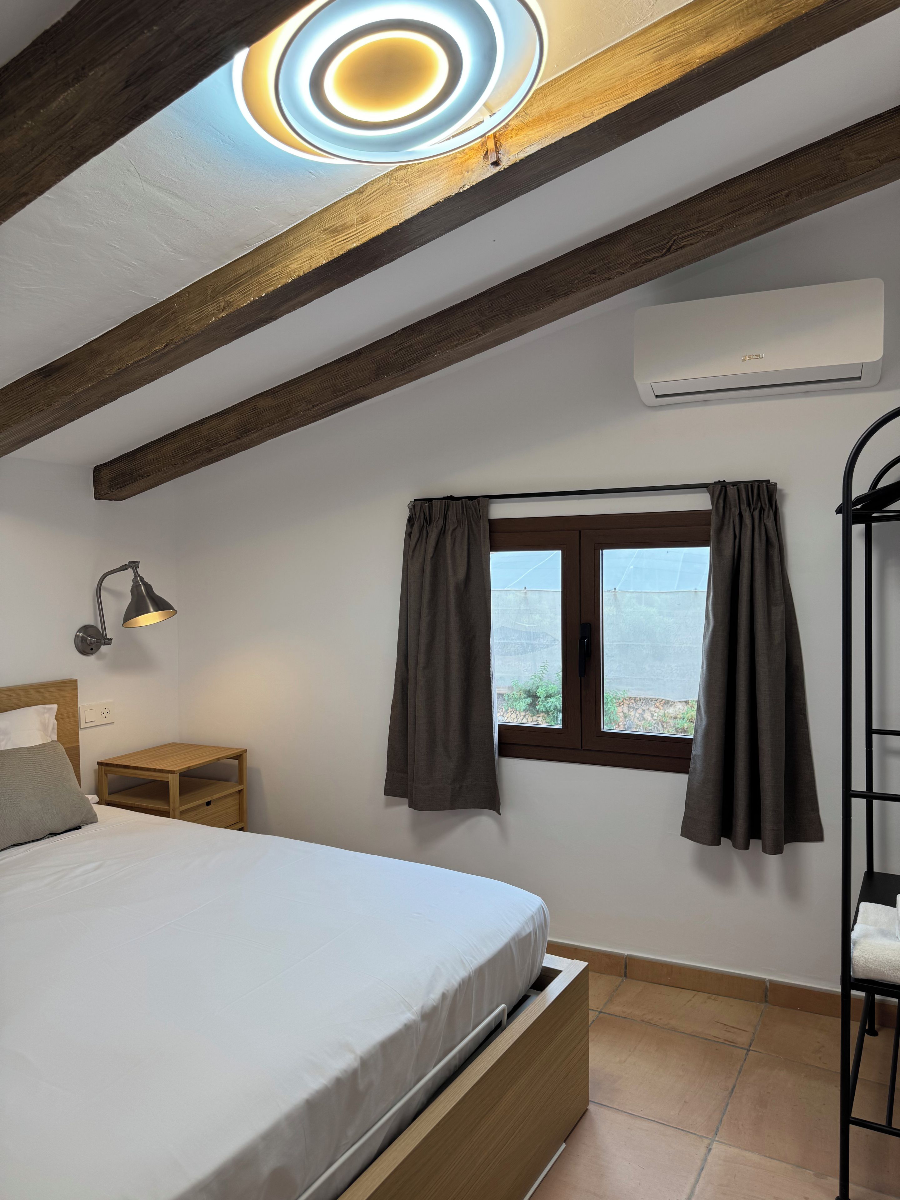 Un dormitorio con cama, lámpara de noche, ventana con cortinas, vigas de madera y unidad de aire acondicionado.