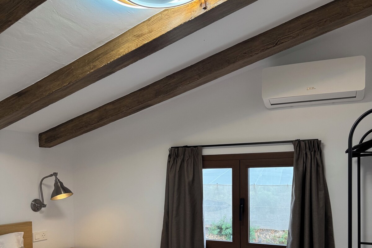 A bedroom with a bed, bedside lamp, window with curtains, wooden beams, and an air conditioning unit.