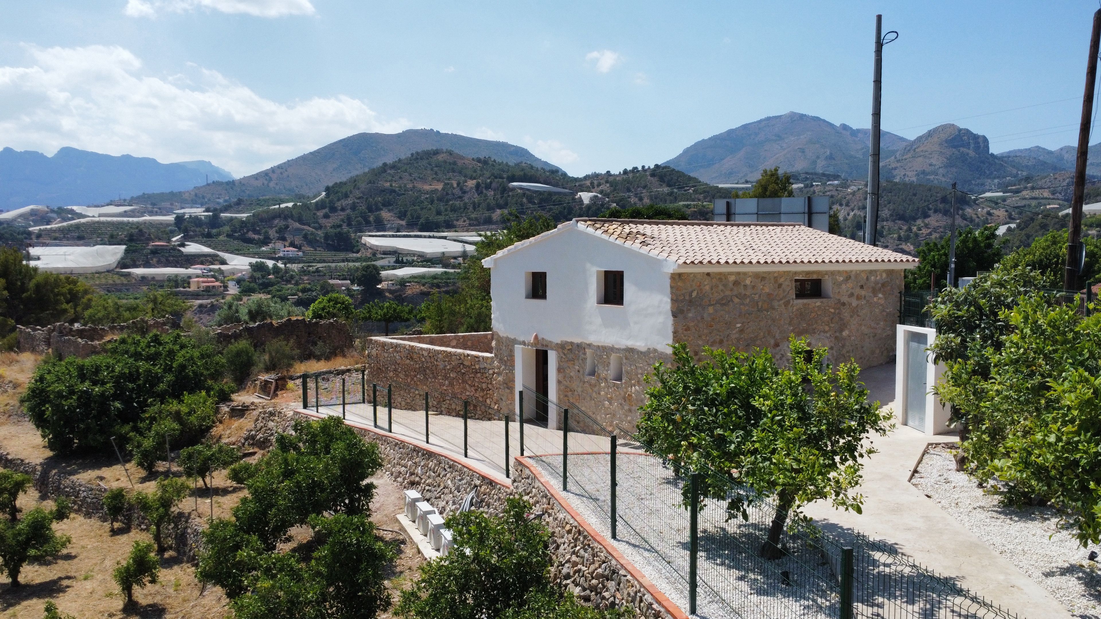 A stone building with a tiled roof, surrounded by a fence and greenery, overlooking a mountainous landscape.