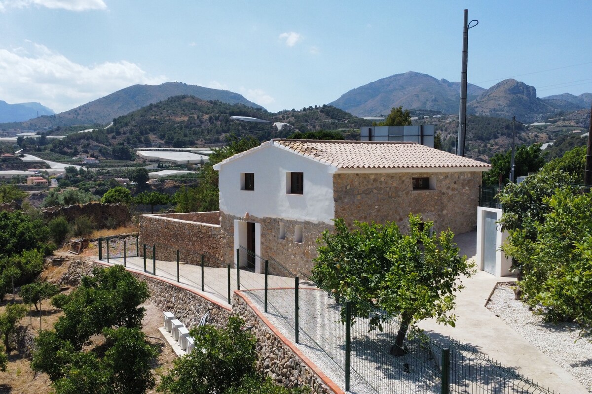 Un edificio de piedra con techo de tejas, rodeado de una valla y vegetación, con vistas a un paisaje montañoso.