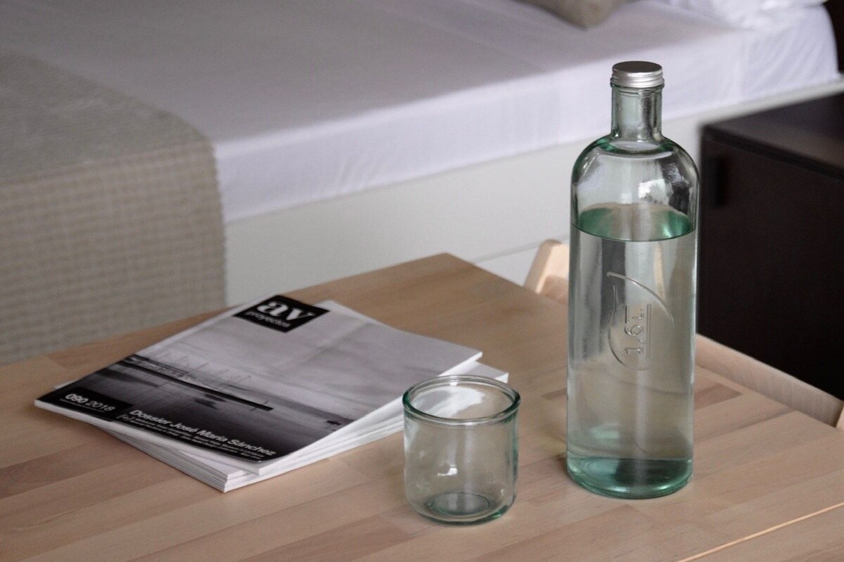 Una cama bien hecha con almohadas, una botella de vidrio y una revista sobre una mesa de madera en una habitación de hotel.